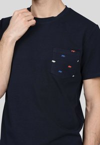 T-shirt en coton bleu marine avec une poche poitrine ornée d'un motif de petites silhouettes d'animaux blanches, bleues et rouges. Modèle à manches courtes.