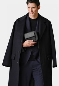 Black leather bag with a fold-over flap held by a person in a dark wool coat over a matching suit, featuring a notched collar and smooth texture.