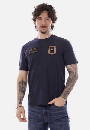 Uomo con capelli ricci che indossa una maglietta blu navy con toppe e tatuaggi su entrambe le braccia, in piedi con una mano in tasca.