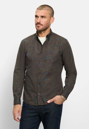 Mann mit kurzen Haaren und Bart, der ein dunkles, gemustertes Hemd mit Knöpfen und dunkle Jeans trägt und mit einer Hand in der Tasche steht.
