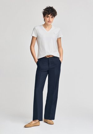 Hvid bomulds V-hals t-shirt, marineblå højtaljede bukser med bælte, og lysebrune loafers, stående mod en neutral baggrund.