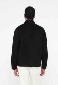 Mann med kort krøllete hår iført svart jakke og hvite bukser, vist bakfra mot en ensfarget hvit bakgrunn.