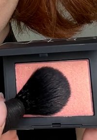 Blush compact with a rectangular shape, soft peach color, and subtle shimmer. A dense black brush is applying the product.