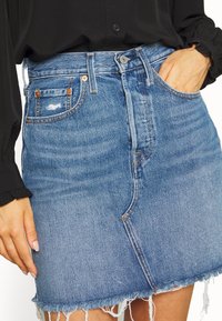 Blå denimkjol med hög midja, fransad nederkant och subtila slitningar, kompletterad med tan-färgade sömmar och fickor.