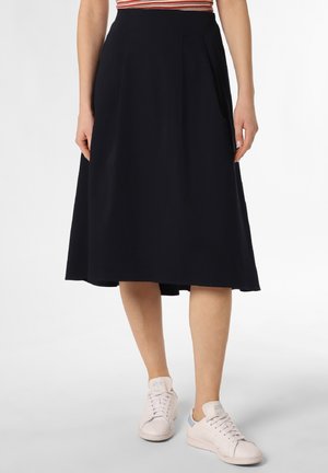 Femme portant une jupe midi noire associée à des baskets blanches, vue de la taille aux pieds sur un fond uni.