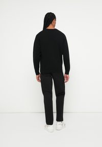Maglione nero a maglia con collo rotondo, abbinato a pantaloni neri e calzature bianche, visto da dietro su uno sfondo bianco semplice.