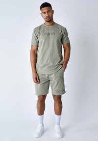 Set di t-shirt e pantaloni corti in cotone verde oliva, con collo tondo, maniche corte e dettaglio del logo ricamato. Indossato con sneakers e calzini bianchi.