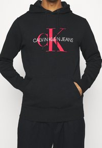 Felpa nera con cappuccio in cotone, con un grande logo grafico rosso e bianco "CALVIN KLEIN JEANS" sul fronte, completa di tasca anteriore.
