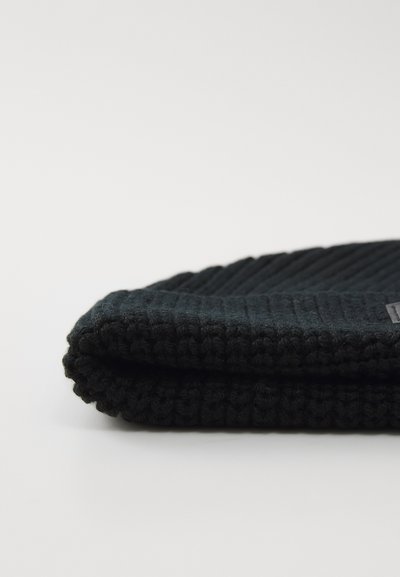 Folded black ribbed knit beanie hat on a white surface, showing texture and a small fabric tag on the side.