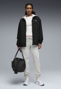 Μαύρο μπουφάν με φερμουάρ πάνω από ένα λευκό πουκάμισο, γκρι φόρμα και λευκά παπούτσια. Κρατά μια μαύρη αθλητική τσάντα. Απλός σχεδιασμός και ουδέτερα χρώματα.
