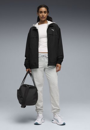 Schwarze Reißverschlussjacke über einem weißen Hemd, graue Jogginghose und weiße Schuhe. Hält eine schwarze Sporttasche. Einfaches Design und neutrale Farben.