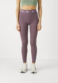 Violet iedarbības legingi ar augstu jostasvīru, uz kuriem ir gaiši pelēks Adidas logo un šuvju detaļas. Valkāti ar baltām sporta kurpēm.