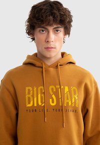 Hnědá bavlněná mikina s kapucí s textem "BIG STAR" v žluté barvě, se stahovací šňůrkou a měkkou strukturou. Jednoduchý, moderní design.