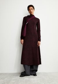 Langes, enganliegendes Kleid in dunkelviolett mit einem gewellten schwarzen Muster, kombiniert mit weiten grauen Hosen. Ein passender lila Riemen überquert den Körper.