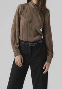 Femme portant un chemisier long à manches pleats marron rentré dans un pantalon noir taille haute avec une ceinture noire et une boucle en argent.