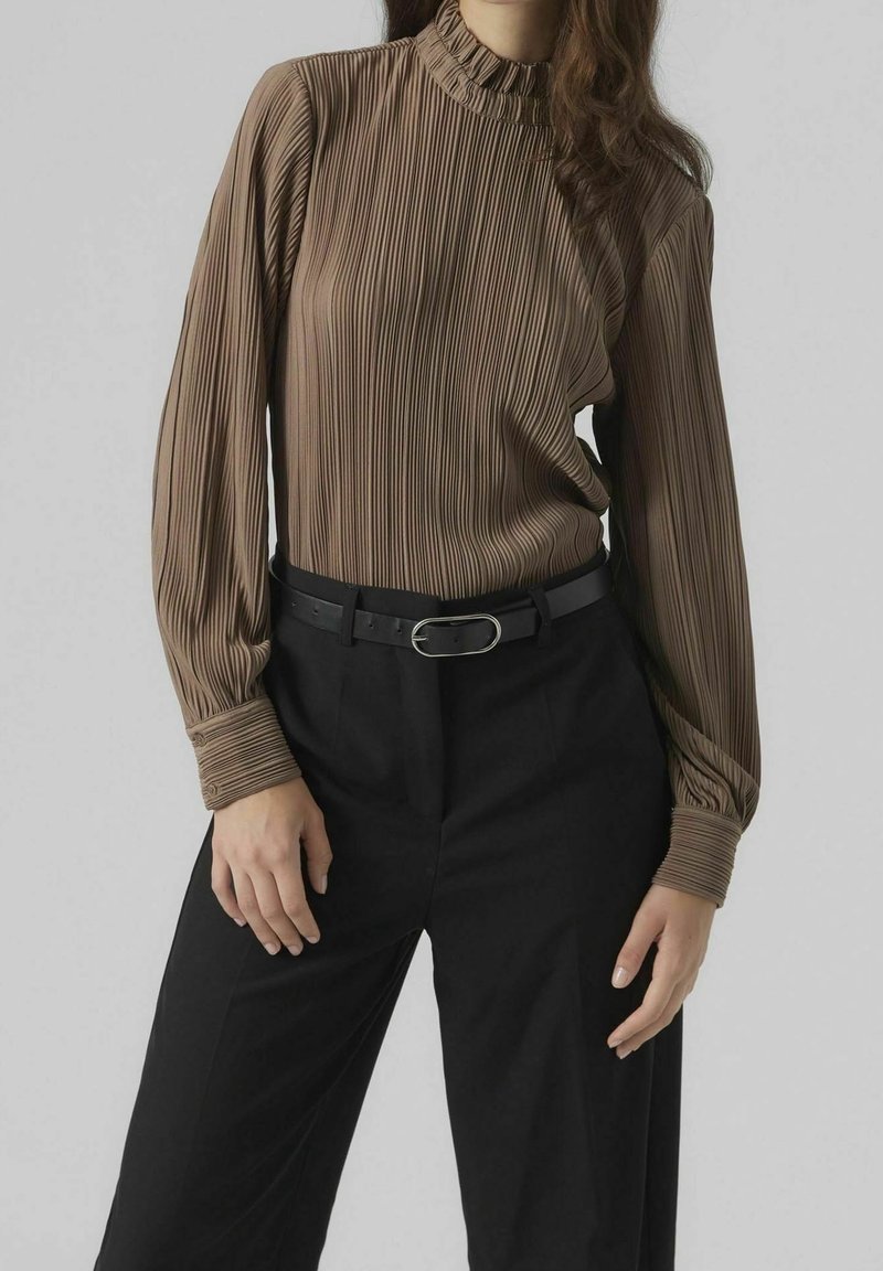 Femme portant un chemisier long à manches pleats marron rentré dans un pantalon noir taille haute avec une ceinture noire et une boucle en argent.