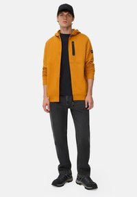 Jeune homme portant un hoodie zippé moutarde, une casquette noire, un t-shirt noir, un jean gris foncé et des baskets noires, se tenant devant un fond blanc.