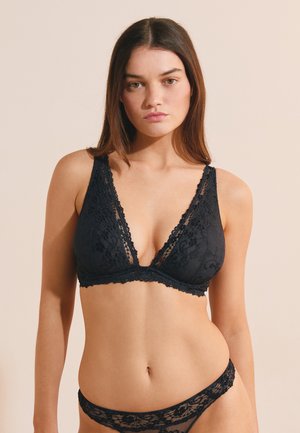Joven mujer con bralette de encaje negro y ropa interior de encaje a juego, de pie frente a un fondo beige.