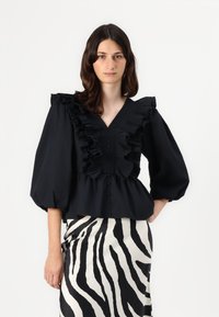Svart bluse med voluminøse flared ermer, dyp v-hals, frontknapper og peplumliv, paret med et svart-hvitt zebra-mønstret skjørt.