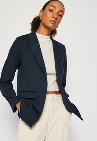 Veste navy à la coupe ajustée, fermeture à un bouton, et revers crantés, associée à un haut clair ajusté et à un pantalon beige texturé.