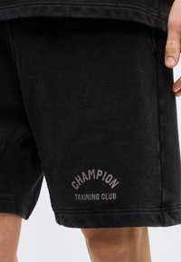 Μαύροι σορτς με την επιγραφή "Champion Training Club" στο αριστερό μηρό, φορεμένα από ένα άτομο που εμφανίζεται από το μέσο του μηρού έως τη μέση.