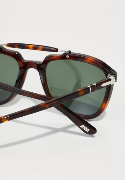 Τετράγωνα γυαλιά ηλίου με καφέ καβουρίσια σχέδια και πράσινους φακούς, μεταλλικές λεπτομέρειες στο σκελετό και κομψά καφέ χερούλια που διασταυρώνονται στο κέντρο.