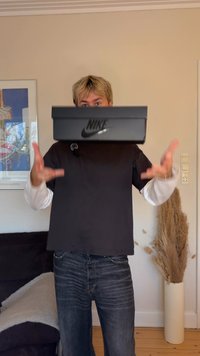 Caixa de sapatos preta da Nike com logótipo, segurada à frente de uma pessoa vestindo uma t-shirt escura e mangas compridas claras, em pé dentro de um espaço com decoração neutra.