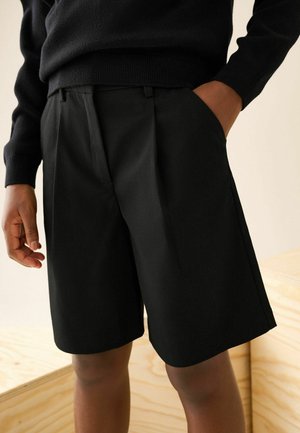 Personne portant un short noir sur mesure à hauteur du genou et un pull noir avec une main dans la poche, sur un fond en bois clair
