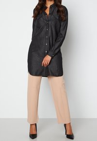 Svart denim skjortklänning med två bröstfickor, långa ärmar och sidostrimlor, kombinerad med beige skräddarsydda byxor och svarta klackskor.