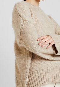 Pull en tricot beige à coupe décontractée, doté de poignets et d'un ourlet côtelés, d'un tissu texturé et de manches raglan pour une apparence douce.