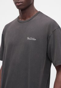 Άνδρας που φορά σκοτεινό γκρι t-shirt Hollister με κοντά μανίκια και στρογγυλή λαιμόκοψη σε απλό φωτεινό φόντο.