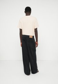 T-shirt de algodão bege e calças largas pretas com riscas finas e um emblema em pele na cintura, vistas por trás.