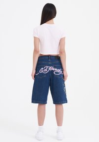 Camiseta corta rosa combinada con bermudas de denim azules que presentan texto bordado en blanco "ED Hardy" y diseños florales en la parte trasera.
