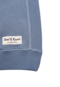 Sininen puuvillaneule, jossa on ribbackit hihansuissa ja helmassa. Tässä on valkoinen etiketti, jossa on tekstit "Band of Rascals" ja "Finest Organic Kids Clothes".