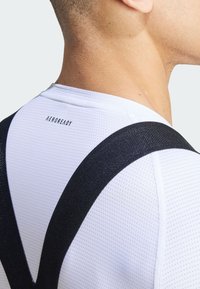 Weißes AEROREADY-Technikshirt mit schwarzen Trägern. Verfügt über ein strukturiertes Muster und ein Logo am hinteren Halsbereich. Nahaufnahme der Schultern.