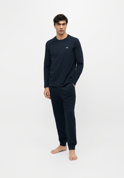 Homme debout pieds nus, portant une chemise noire à manches longues et un pantalon assorti sur un fond blanc uni.