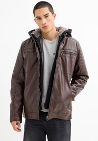 Chaqueta de cuero marrón con capucha, forro de felpa gris, cierre delantero con cremallera y detalles acolchados en los hombros. Lleva puesta una camiseta gris.