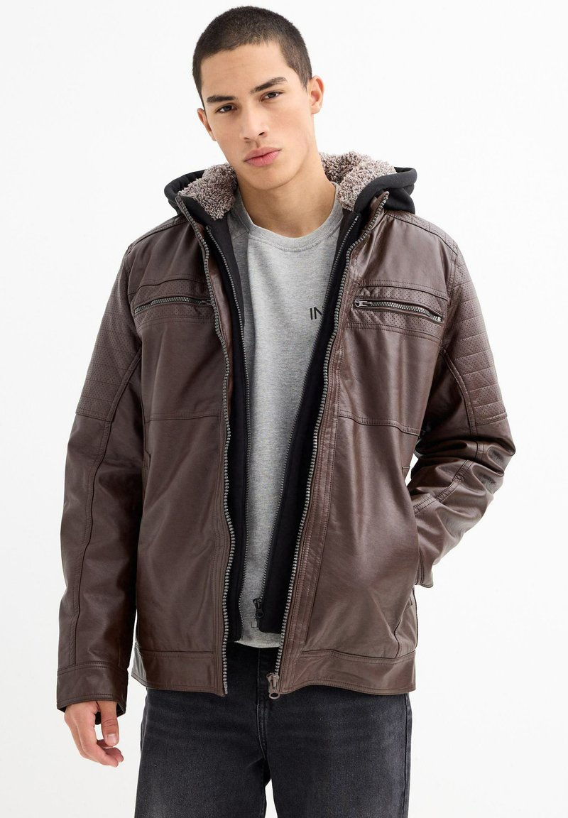 Chaqueta de cuero marrón con capucha, forro de felpa gris, cierre delantero con cremallera y detalles acolchados en los hombros. Lleva puesta una camiseta gris.