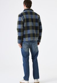 Mann, der mit dem Rücken zur Kamera steht, trägt eine blau-grün karierte Jacke, blaue Jeans und weiße Sneaker vor einem schlichten weißen Hintergrund.