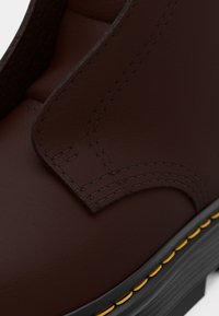 Bota de cuero marrón con textura suave, costura amarilla a lo largo de la suela y un diseño sutil con un detalle de costura reforzada.