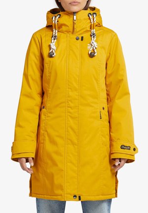 Femme portant un long manteau d'hiver jaune à capuche avec cordons blancs, fermeture éclair devant et poignets boutonnés, associé à un jean bleu.