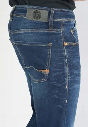 Jean en denim bleu foncé avec une finition texturée, des coutures visibles, une poche arrière avec un accent beige et une étiquette de logo sur la taille.