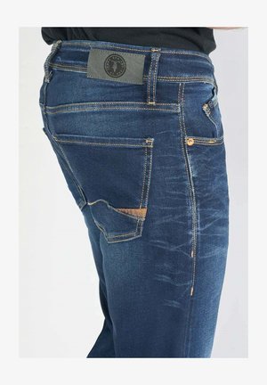 Jean en denim bleu foncé avec une finition texturée, des coutures visibles, une poche arrière avec un accent beige et une étiquette de logo sur la taille.