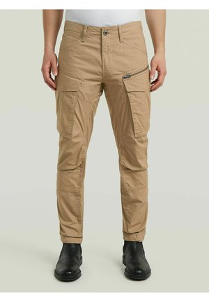 Pantaloni cargo beige con diverse tasche, tra cui una con zip sulla coscia destra, indossati con stivali neri in pelle e una maglia bianca.