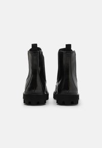 Botas de tobillo de charol negro con gruesas suelas de goma, paneles laterales elásticos y tiradores en la parte trasera para facilitar su uso.