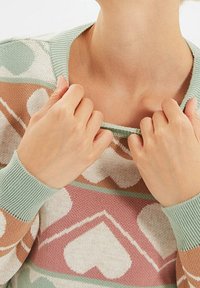 Person som justerar halsringningen på en mönstrad tröja med stora hjärtformer i pastellgröna, rosa och beige ton.