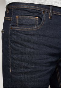Donkerblauwe denim jeans hebben contrasterende oranje stiksels, vijf zakken en bronzen hardware accenten aan de voorkant en achterkant.