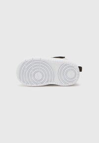 Semelle de sneaker blanche avec des motifs de traction circulaires et des rainures. Présente une sangle noire sur le côté. Matériau en caoutchouc lisse.