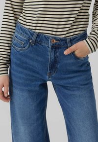 Pantalones vaqueros de mezclilla azul con cintura alta, bolsillos frontales y detalle de botón de cobre, combinados con una camisa de manga larga a rayas en blanco y negro.