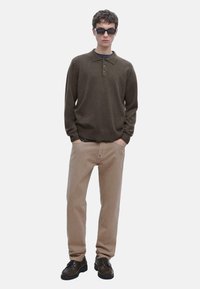 Giovane uomo che indossa un polo a maniche lunghe marrone scuro, pantaloni beige, scarpe nere e occhiali da sole rettangolari neri, in piedi con le mani in tasca.